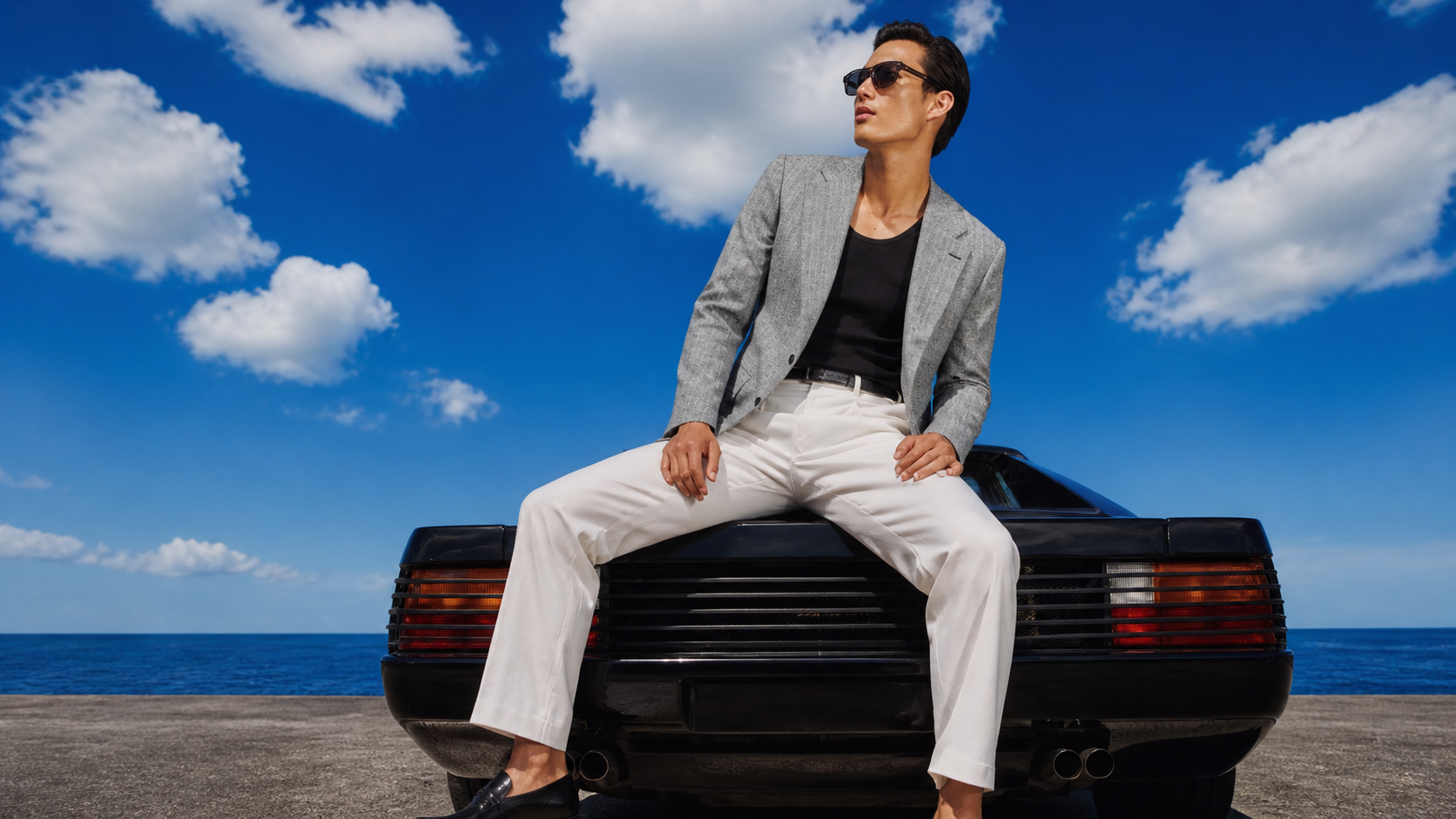 Man in a patterned grey blazer sitting on a black sports car under a blue sky